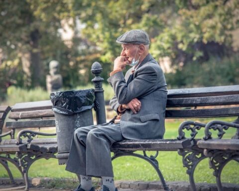 Elderly man in a gray suit and flat cap sitting alone on a park bench with a face mask pulled down, appearing thoughtful or concerned.