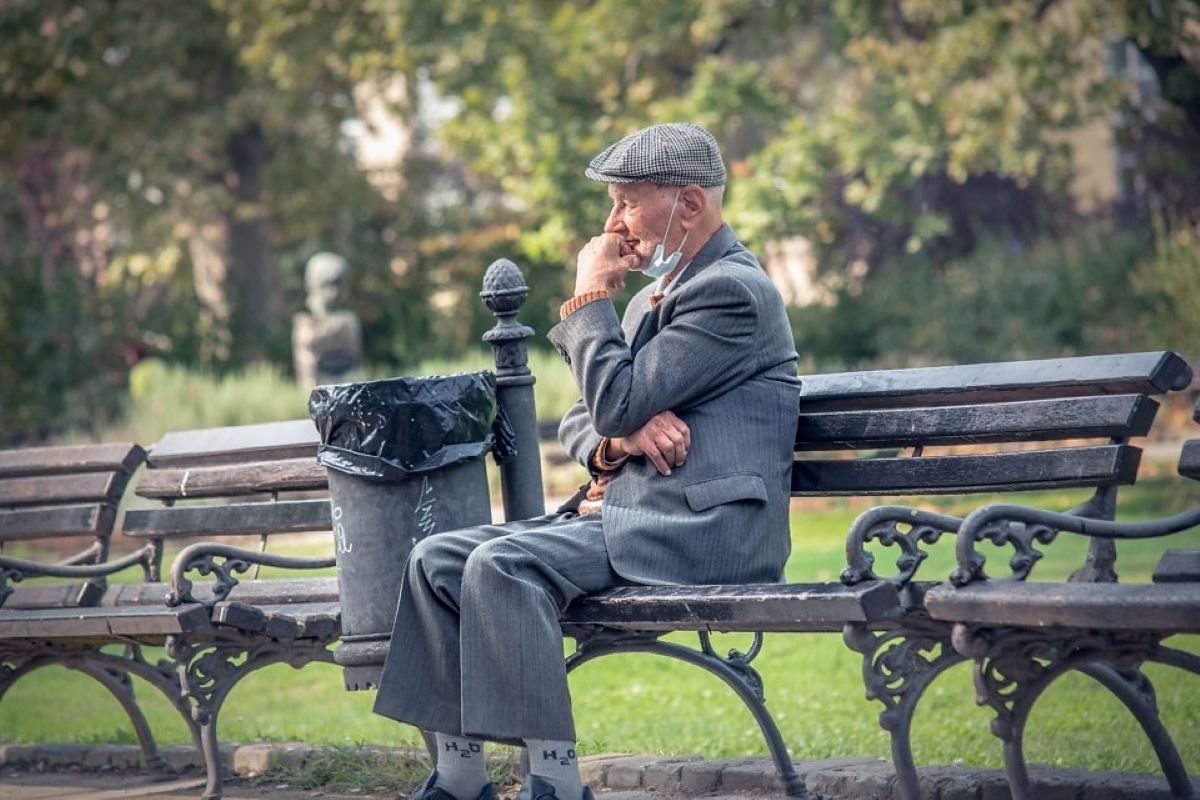 Elderly man in a gray suit and flat cap sitting alone on a park bench with a face mask pulled down, appearing thoughtful or concerned.