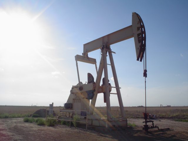 An oil pumpjack (nodding donkey) extracting crude oil in a desert landscape, with the mechanical arm mid-cycle against a clear sky.
