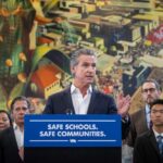 Governor Gavin Newsom speaking at a podium with "SAFE SCHOOLS. SAFE COMMUNITIES." banner, surrounded by lawmakers and community members during the immigration protection laws signing ceremony in Los Angeles.