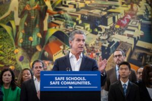 Governor Gavin Newsom speaking at a podium with "SAFE SCHOOLS. SAFE COMMUNITIES." banner, surrounded by lawmakers and community members during the immigration protection laws signing ceremony in Los Angeles.