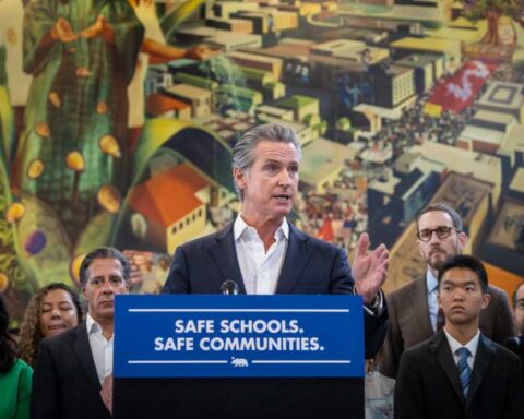 Governor Gavin Newsom speaking at a podium with "SAFE SCHOOLS. SAFE COMMUNITIES." banner, surrounded by lawmakers and community members during the immigration protection laws signing ceremony in Los Angeles.
