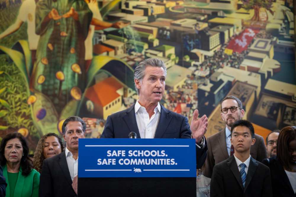 Governor Gavin Newsom speaking at a podium with "SAFE SCHOOLS. SAFE COMMUNITIES." banner, surrounded by lawmakers and community members during the immigration protection laws signing ceremony in Los Angeles.