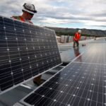 Workers installing solar panels on a commercial rooftop with mountains visible in the background.