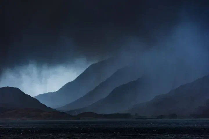 Dramatic rainfall falling over mountainous terrain with dark storm clouds, showing intense precipitation sweeping across a Scottish landscape.
