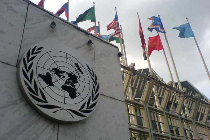 United Nations Building in Bangkok showing the UN emblem on a white marble wall alongside multiple national flags flying on poles, representing international cooperation on global issues.