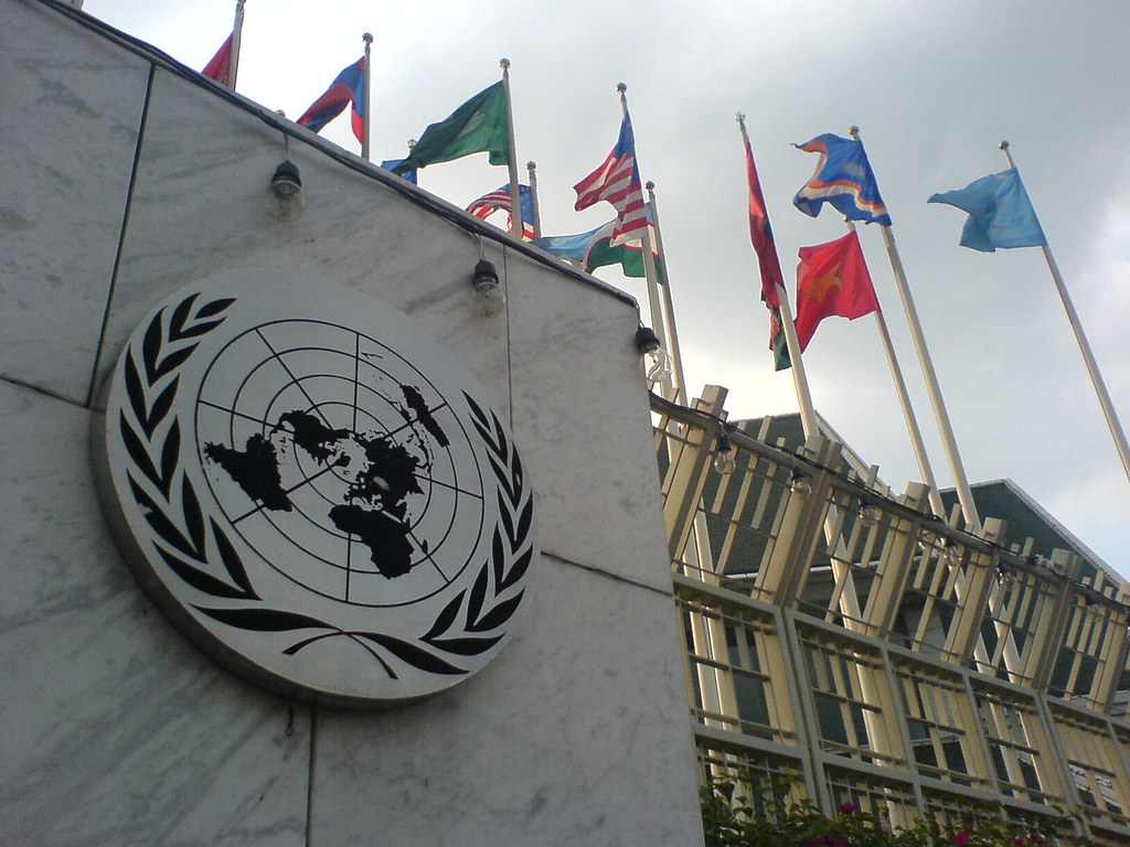 United Nations Building in Bangkok showing the UN emblem on a white marble wall alongside multiple national flags flying on poles, representing international cooperation on global issues.