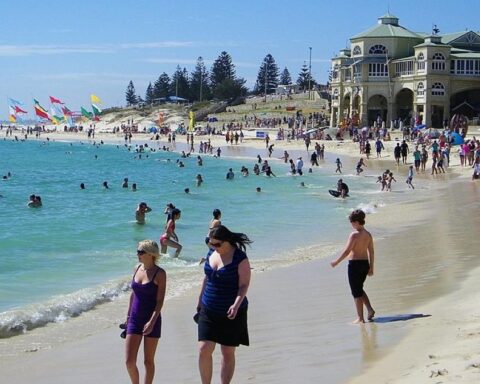 Crowded summer scene at Cottesloe Beach in Perth, Western Australia, with people swimming and relaxing on the sand near the heritage-style Cottesloe Pavilion.