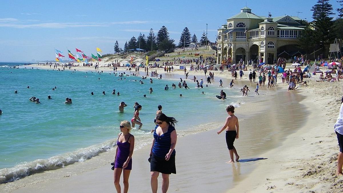 Crowded summer scene at Cottesloe Beach in Perth, Western Australia, with people swimming and relaxing on the sand near the heritage-style Cottesloe Pavilion.
