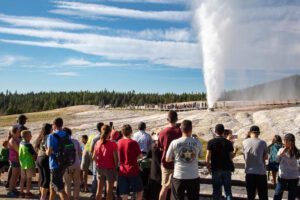 Visitors gathered around Beehive Geyser in Yellowstone National Park during a powerful eruption, with steam and water shooting into the sky.