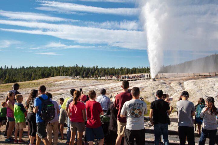 Visitors gathered around Beehive Geyser in Yellowstone National Park during a powerful eruption, with steam and water shooting into the sky.