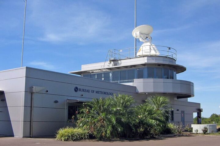 The Bureau of Meteorology’s Darwin Airport Met Office building featuring a white radar dome, rooftop antennas and surrounding palm trees under a clear blue sky