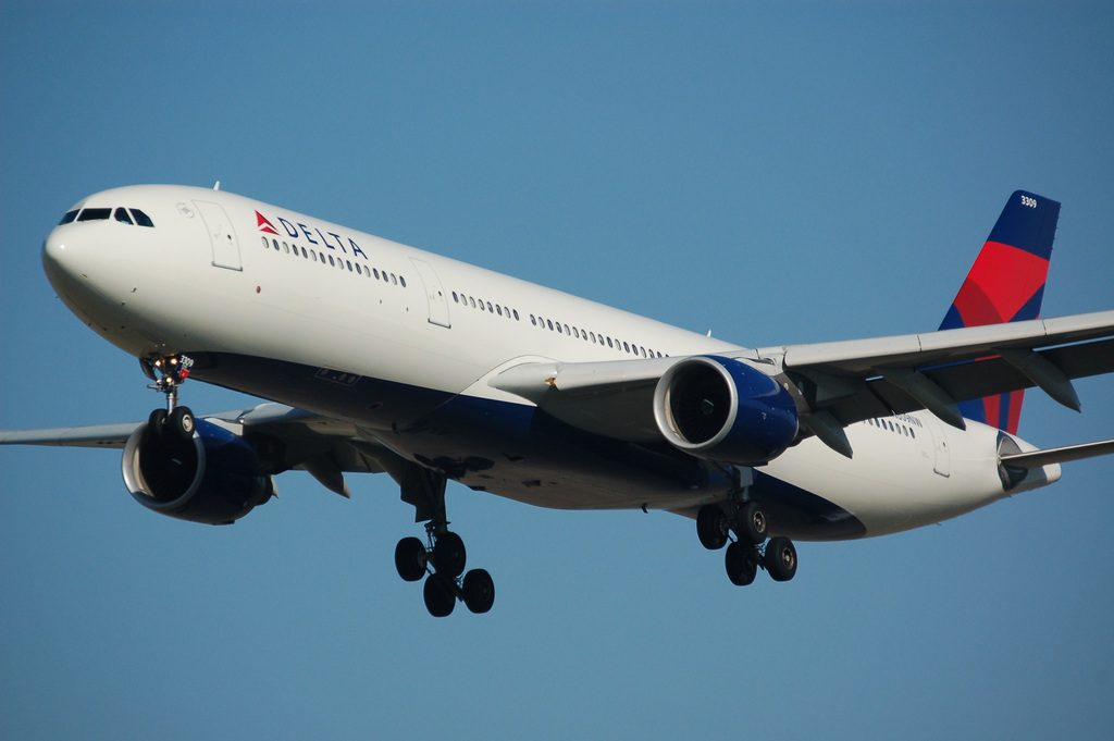 Delta Air Lines passenger aircraft with white fuselage and blue engines preparing to land with landing gear deployed against a blue sky background.