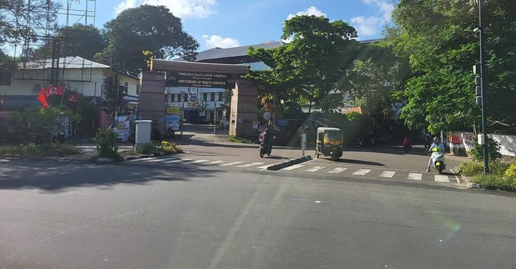 Front view of Directorate of Health Services building in Kerala with trees, vehicles, and clear sky.