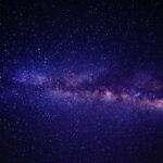 A stunning night sky photograph showing the Milky Way galaxy with its bright central band of stars and cosmic dust clouds stretching across a deep blue-purple background filled with countless stars.
