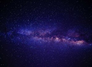 A stunning night sky photograph showing the Milky Way galaxy with its bright central band of stars and cosmic dust clouds stretching across a deep blue-purple background filled with countless stars.