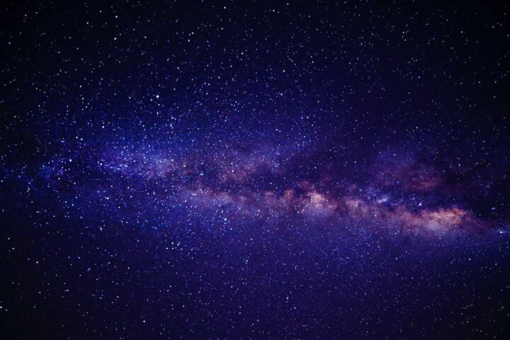 A stunning night sky photograph showing the Milky Way galaxy with its bright central band of stars and cosmic dust clouds stretching across a deep blue-purple background filled with countless stars.