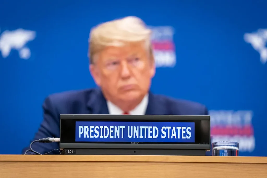 Donald Trump blurred in background with a conference placard reading “President United States” in focus, captured at an international session.