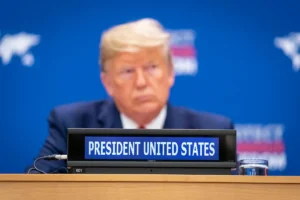 Donald Trump blurred in background with a conference placard reading “President United States” in focus, captured at an international session.
