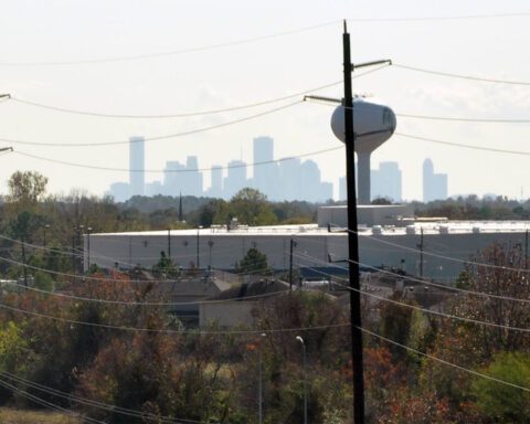 Distant view of downtown Houston skyline with utility poles and water tower in the foreground, under slightly hazy skies, symbolizing the city’s infrastructure and energy network.