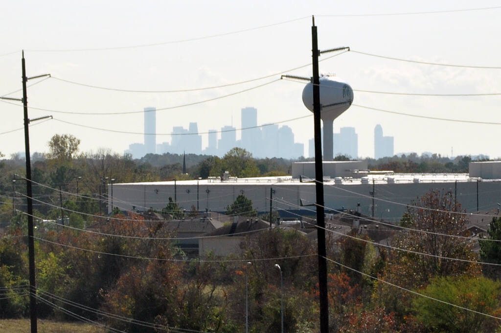 Distant view of downtown Houston skyline with utility poles and water tower in the foreground, under slightly hazy skies, symbolizing the city’s infrastructure and energy network.