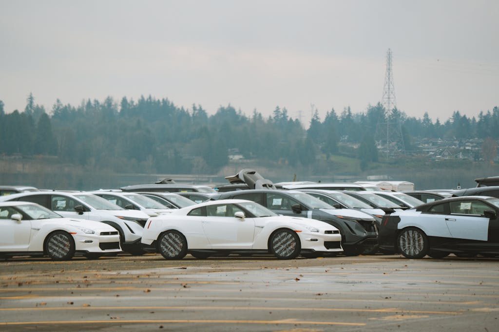 A row of white Nissan GT-R sports cars parked in an outdoor storage lot with forested hills and a transmission tower visible in the background under an overcast sky.