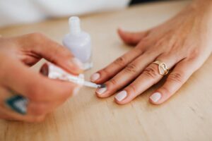 Close-up of a manicurist applying white nail polish to a woman's hand wearing a gold heart-shaped ring on a wooden table surface.