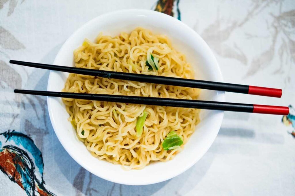 Bowl of yellow ramen noodles with small green garnish served in a white round bowl with black chopsticks that have red tips resting across the top
