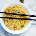 Bowl of yellow ramen noodles with small green garnish served in a white round bowl with black chopsticks that have red tips resting across the top
