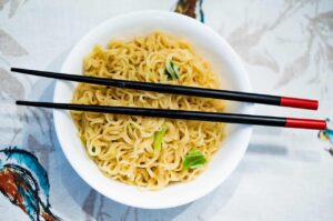 Bowl of yellow ramen noodles with small green garnish served in a white round bowl with black chopsticks that have red tips resting across the top