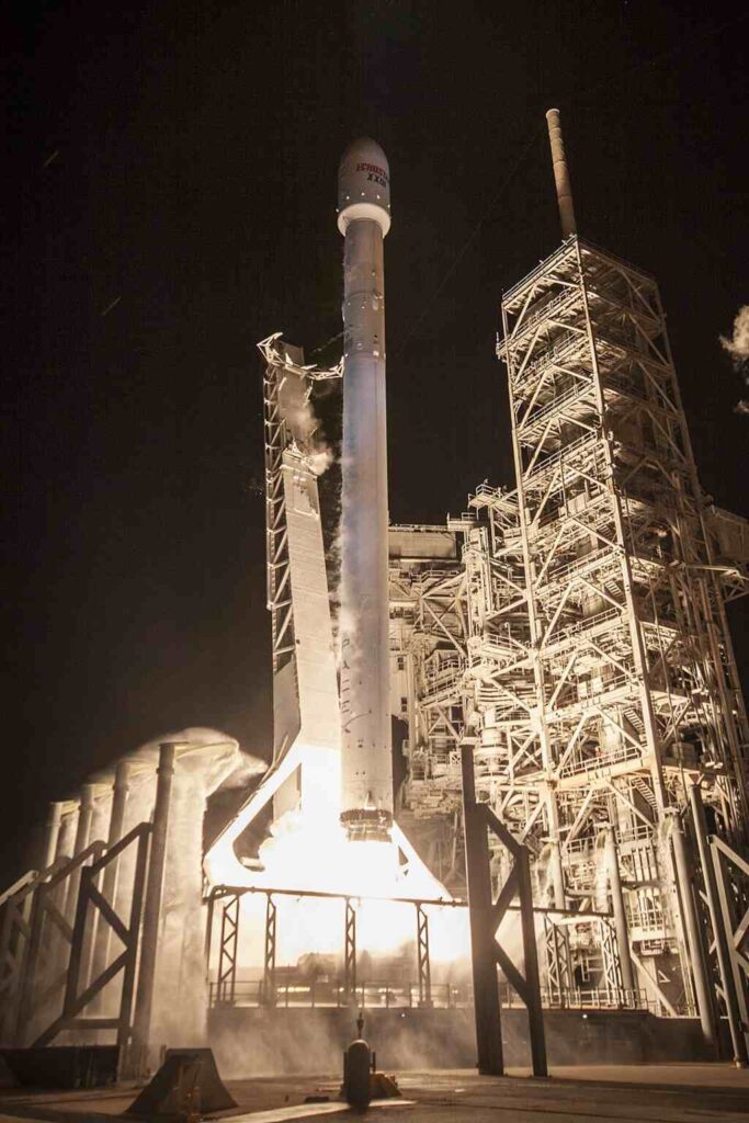 A SpaceX Falcon 9 rocket launches the EchoStar XXIII satellite into a clear blue sky, with intense exhaust plume visible.