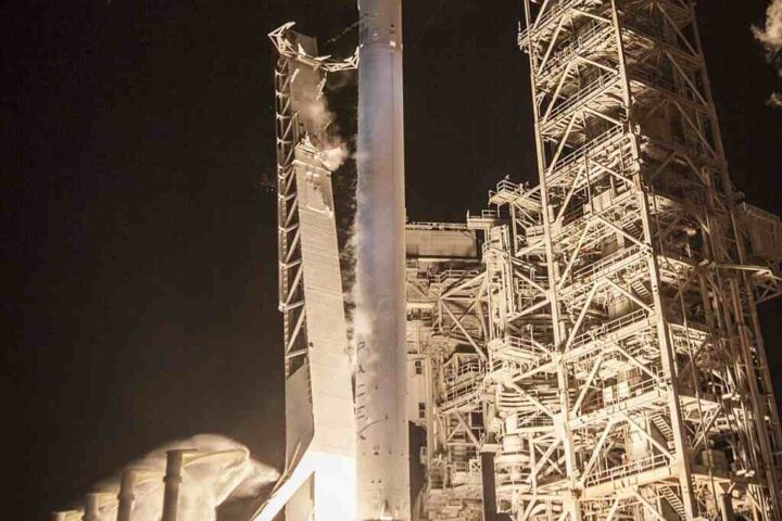 A SpaceX Falcon 9 rocket launches the EchoStar XXIII satellite into a clear blue sky, with intense exhaust plume visible.