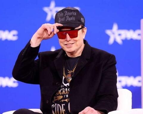 Elon Musk wearing sunglasses and a cap, speaking on stage at the 2025 Conservative Political Action Conference in Maryland.