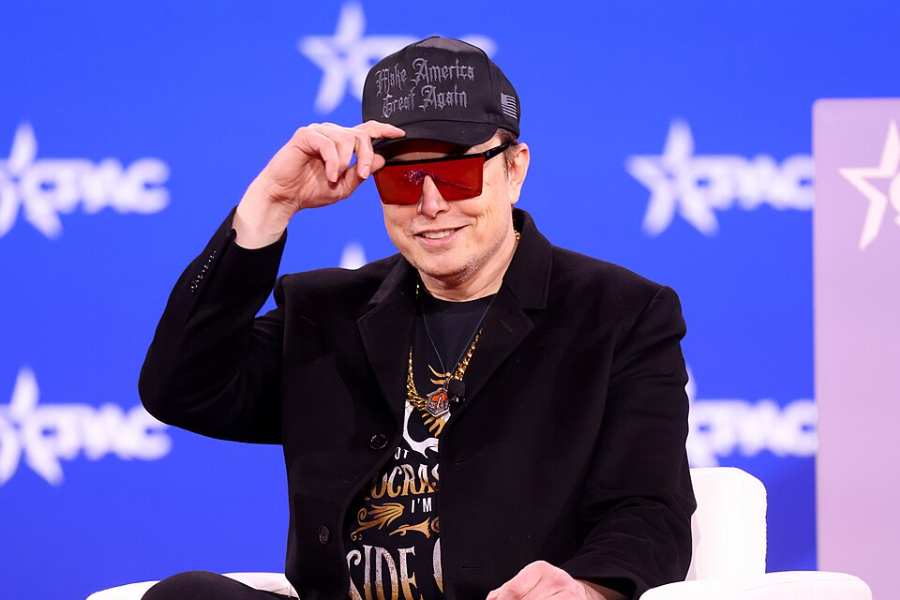 Elon Musk wearing sunglasses and a cap, speaking on stage at the 2025 Conservative Political Action Conference in Maryland.