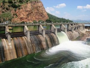 A concrete dam with multiple water release gates showing water flowing through spillways, set against a backdrop of rocky cliffs and mountains.