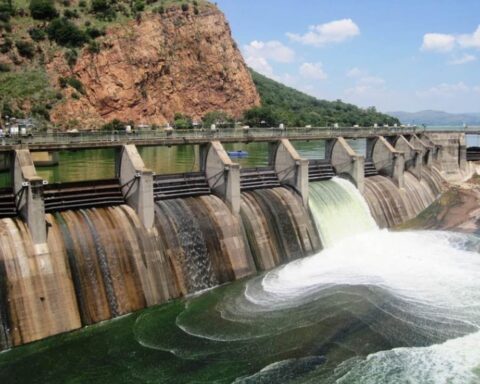 A concrete dam with multiple water release gates showing water flowing through spillways, set against a backdrop of rocky cliffs and mountains.