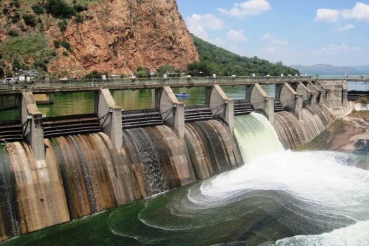 A concrete dam with multiple water release gates showing water flowing through spillways, set against a backdrop of rocky cliffs and mountains.