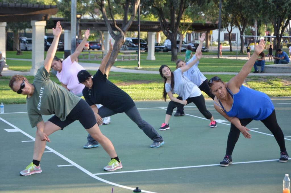 Diverse group of people practicing yoga or stretching exercises outdoors in a park, with five individuals in various side bend poses on a sports court.