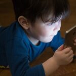 Young boy in blue shirt looking down at smartphone screen while sitting at wooden table