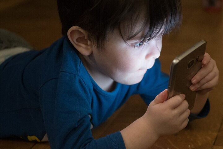 Young boy in blue shirt looking down at smartphone screen while sitting at wooden table