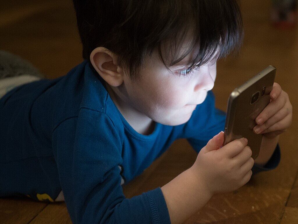 Young boy in blue shirt looking down at smartphone screen while sitting at wooden table