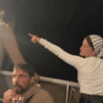 Nighttime scene aboard a Gaza aid vessel showing activists in light-colored clothing on deck, with one woman wearing a checkered headscarf pointing upward at unseen surveillance drones while others look on with concern.