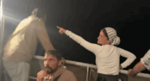 Nighttime scene aboard a Gaza aid vessel showing activists in light-colored clothing on deck, with one woman wearing a checkered headscarf pointing upward at unseen surveillance drones while others look on with concern.