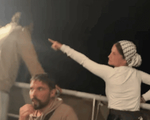 Nighttime scene aboard a Gaza aid vessel showing activists in light-colored clothing on deck, with one woman wearing a checkered headscarf pointing upward at unseen surveillance drones while others look on with concern.