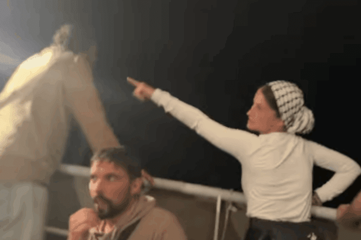Nighttime scene aboard a Gaza aid vessel showing activists in light-colored clothing on deck, with one woman wearing a checkered headscarf pointing upward at unseen surveillance drones while others look on with concern.