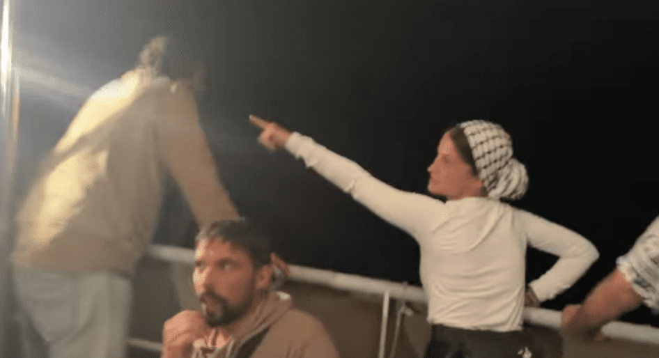 Nighttime scene aboard a Gaza aid vessel showing activists in light-colored clothing on deck, with one woman wearing a checkered headscarf pointing upward at unseen surveillance drones while others look on with concern.