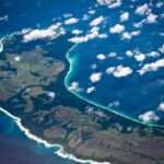 An aerial photograph of the lush green, densely forested Teressa Island in the Nicobar archipelago, with a pristine coastline and turquoise waters, illustrating the region's fragile ecological beauty under threat from development.
