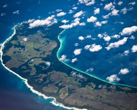An aerial photograph of the lush green, densely forested Teressa Island in the Nicobar archipelago, with a pristine coastline and turquoise waters, illustrating the region's fragile ecological beauty under threat from development.