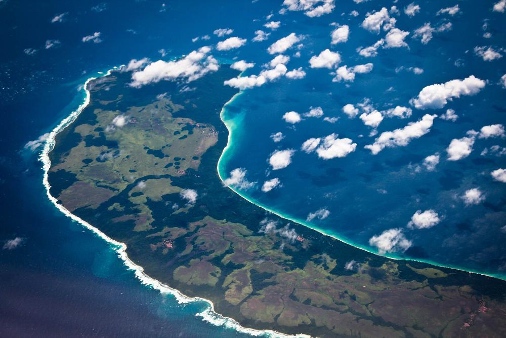 An aerial photograph of the lush green, densely forested Teressa Island in the Nicobar archipelago, with a pristine coastline and turquoise waters, illustrating the region's fragile ecological beauty under threat from development.