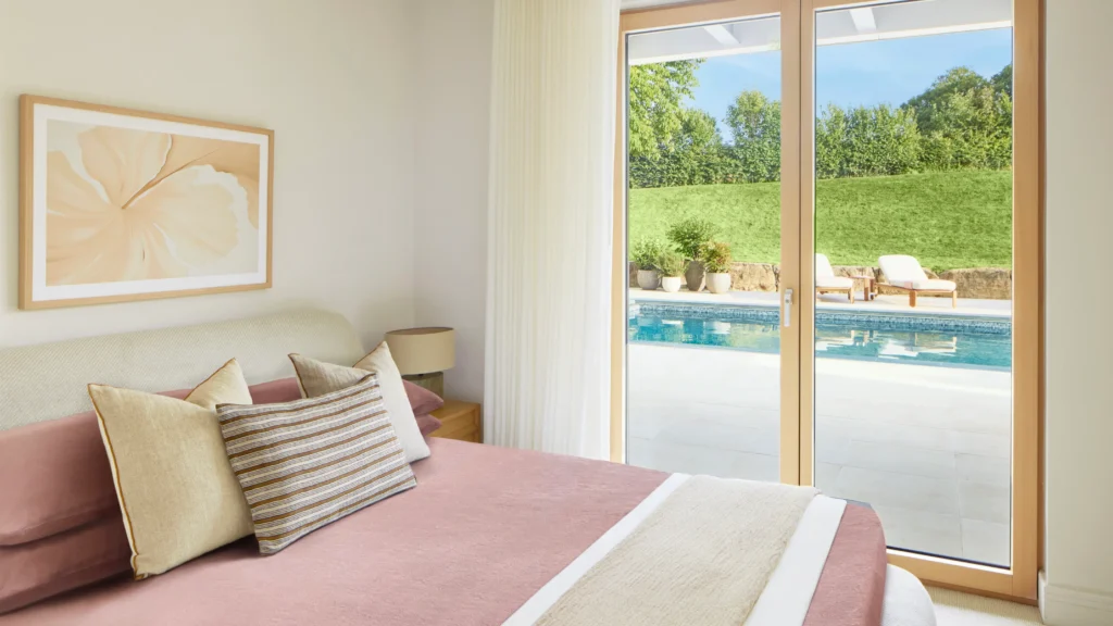 Modern guest bedroom with minimalist decor, natural wood headboard, and floor-to-ceiling glass doors leading to garden area.